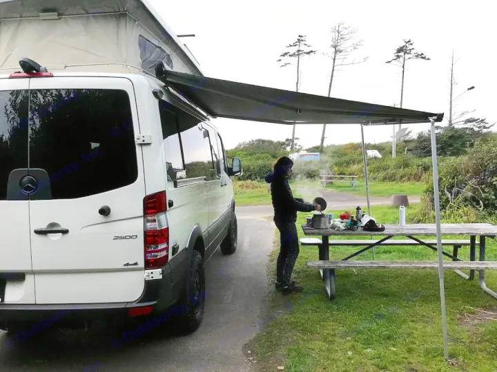 White Sprinter camper van, possibly an RV rental, with an extended awning. A person cooks on a picnic table at a campsite.. Mercedes Sprinter 2018