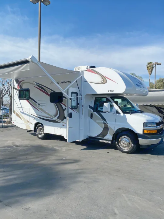 A white Class C motorhome, branded Thor Four Winds, is parked with its awning extended. Ideal for your RV rental needs.. Thor Motor Coach Four Winds 2020