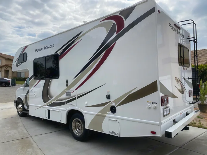 A white Four Winds Class C RV rental, featuring distinctive graphics and a rear ladder, parked on a concrete driveway.. Thor Motor Coach Four Winds 2020