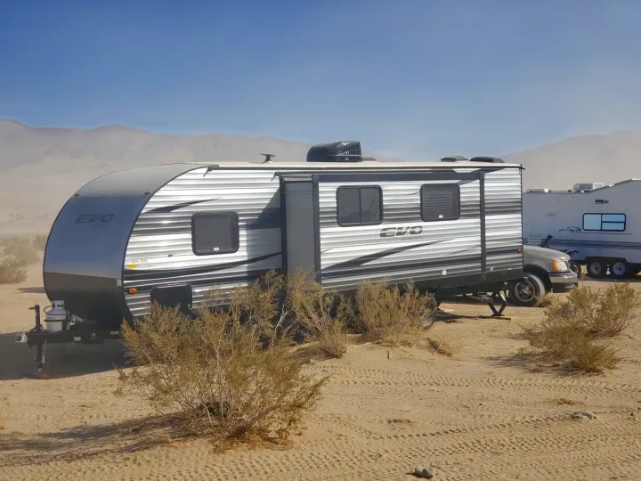 Gray RV rental travel trailer set up in a sandy desert landscape, with a truck, dry bushes, and distant hazy mountains.. Forest River EVO 2020