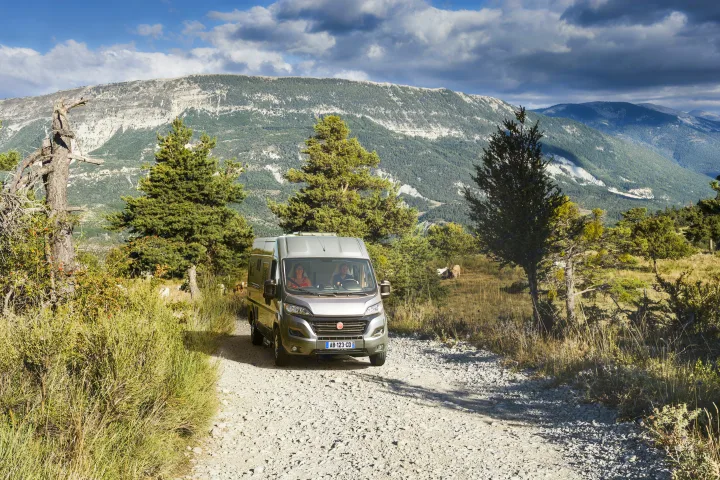 An RV rental with a couple drives a gravel mountain road. Scenic, tree-covered peaks and cloudy skies provide a rugged backdrop.. Pilote v630s 2027
