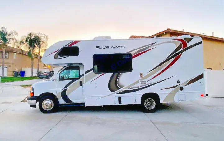 A white Four Winds Class C RV with decorative side stripes is parked on a paved driveway at dusk, ready for rv rental adventures.. Thor Motor Coach Four Winds 2020