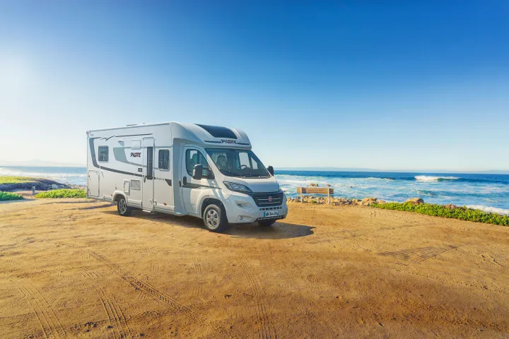 A sleek white RV is parked on a sandy lot by the sunny ocean with waves and a bench, perfect for your next rv rental adventure.. Pilote P746 A 2025