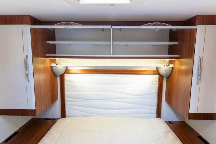 Modern RV rental bedroom interior with open overhead cabinets, a white bed, textured headboard, and integrated lighting.. Pilote P746 A 2025