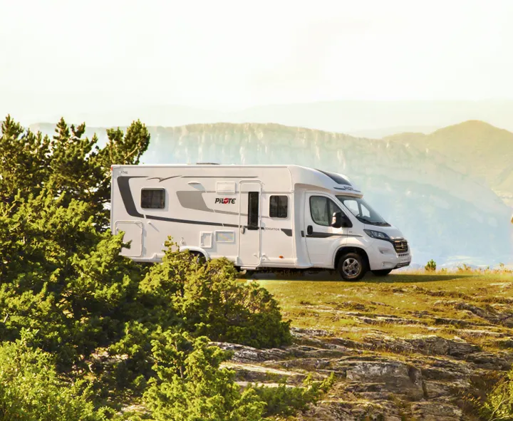 A white Pilote motorhome, suitable for an rv rental, is parked on a rocky cliffside, with hazy mountains in the background.. Pilote P746 A 2025