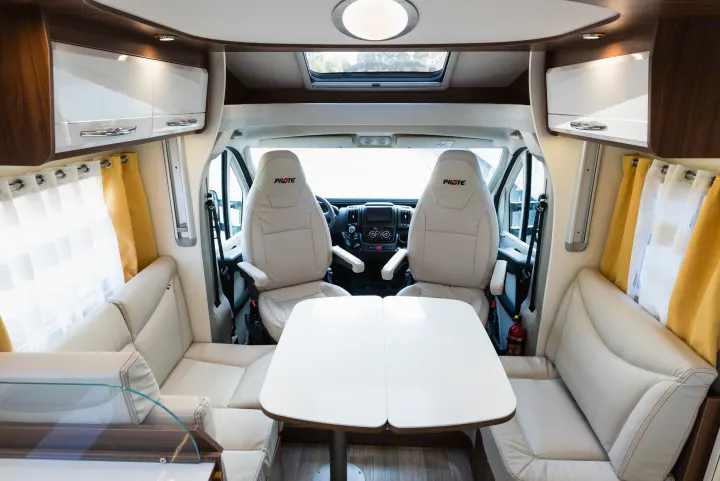 Bright RV rental interior with cream seating, dining table, cockpit view, and overhead storage.. Pilote P746 A 2025