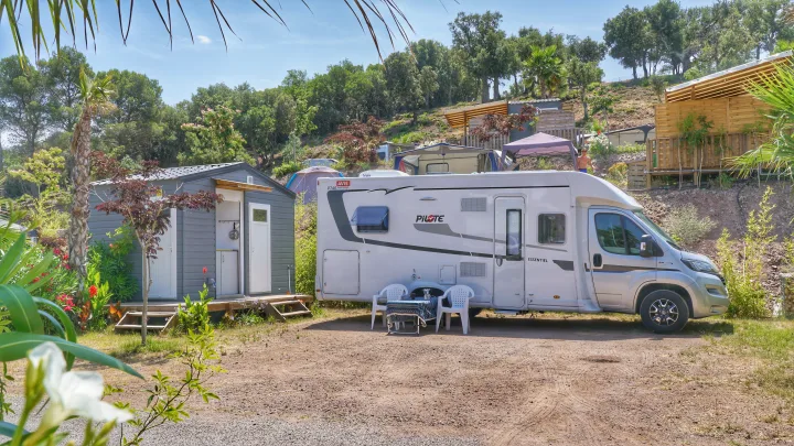 A white RV rental and small gray cabin sit on a dirt pitch at a sunny, tree-lined campsite. Ideal for outdoor adventures.. Pilote P746 A 2025