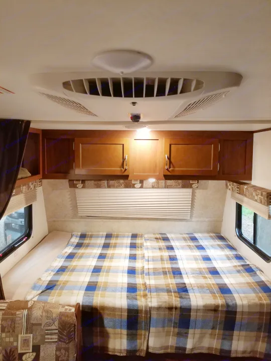 Inside an RV rental, a plaid-covered bed, window, ceiling AC unit, and wooden storage cabinets are visible.. Jayco SLX - 21.5' 2017
