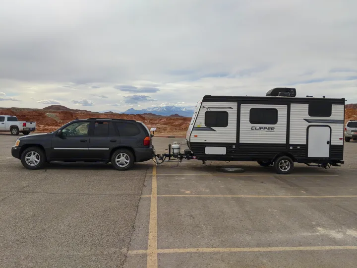 SUV towing an RV rental on an asphalt lot, with red rock formations and snow-capped mountains in the distance.. Forest River Clipper Towable 2020