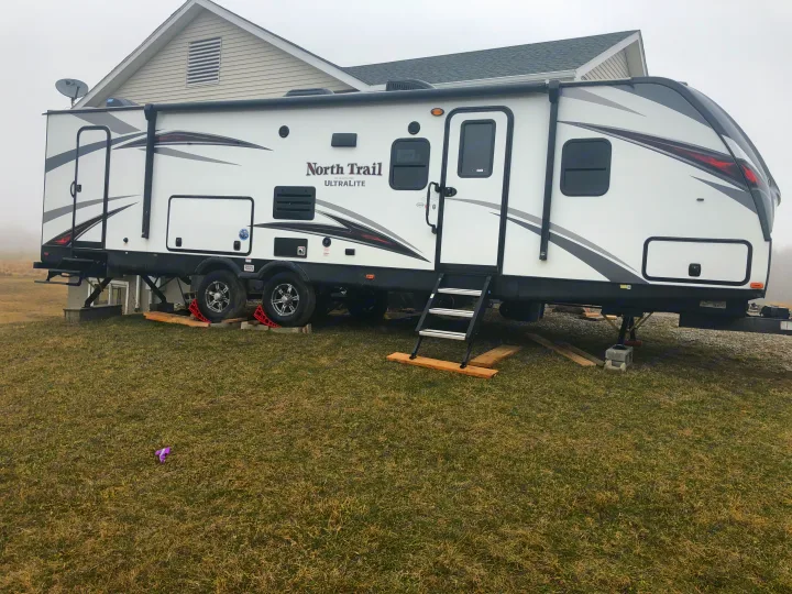 A white North Trail Ultralite RV rental is parked on grass, securely stabilized by jacks and blocks next to a building.. Heartland North Trail 2019