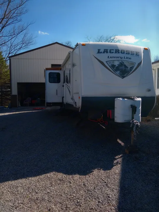 Parked LaCrosse Luxury Lite travel trailer on a gravel lot next to a large shed, ready for RV rental adventures under a clear sky.. Prime Time Lacrosse 2013