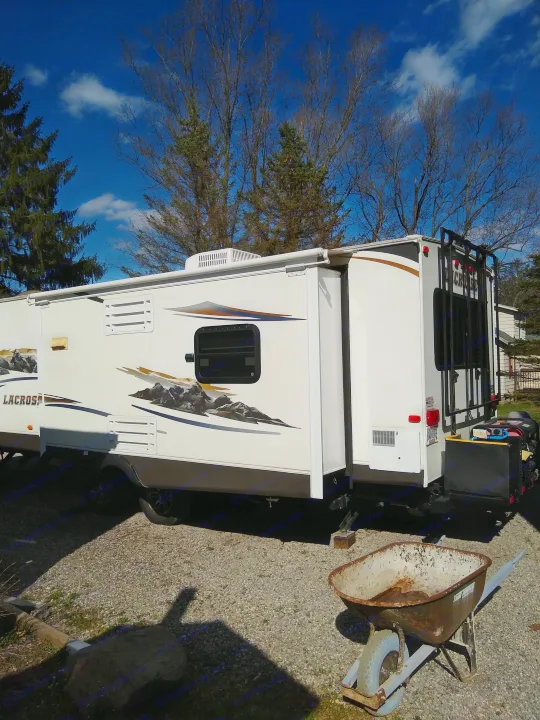 LaCrosse travel trailer RV rental with slide-out extended, parked on gravel; rusty wheelbarrow nearby.. Prime Time Lacrosse 2013