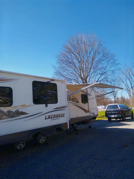 A LaCrosse travel trailer RV rental is set up on gravel with its awning extended. A black pickup truck waits under a clear blue sky.. Prime Time Lacrosse 2013
