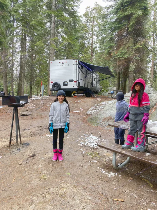 RV rental campsite fun! Kids in warm winter gear explore a muddy, snowy forest next to their white travel trailer, grill, and picnic table.. Keystone Hideout 2020