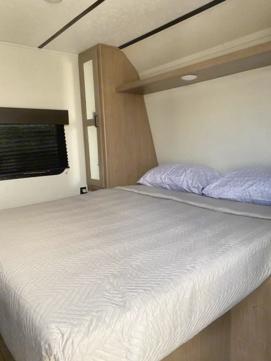 Inviting RV rental bedroom: comfortable bed with chevron comforter, window with black blinds, and built-in storage.. Keystone Hideout 2020