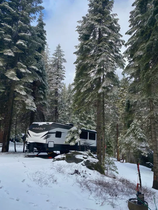 RV rental parked in a tranquil snowy forest campsite. Tall evergreens covered in snow surround the travel trailer.. Keystone Hideout 2020