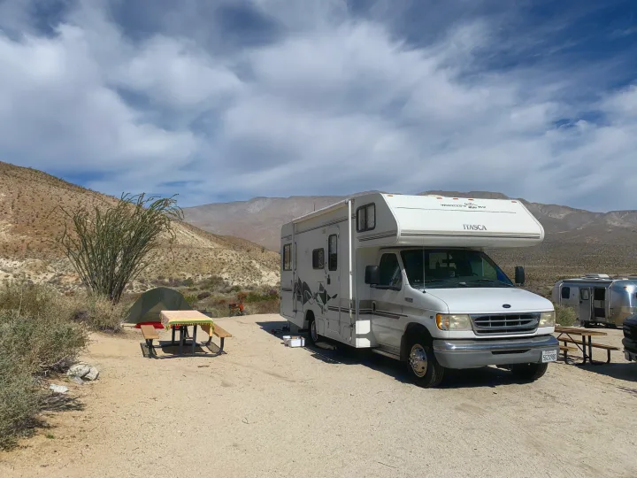 A white Itasca rv rental rests in a dry desert campsite with a tent, picnic table, and Ocotillo plant. Mountains loom under clouds.. Itasca Spirit 1999