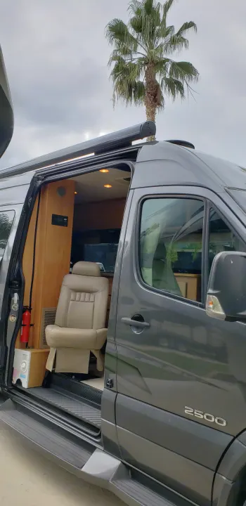 Grey RV rental with open side door, revealing beige interior, chair, and fire extinguisher. Palm tree in background.. ERA Era 2010