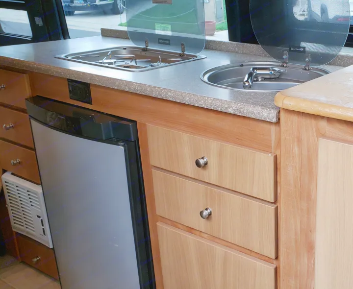 Compact RV rental kitchen with a built-in stove, sink, and refrigerator, featuring light wood cabinets.. ERA Era 2010