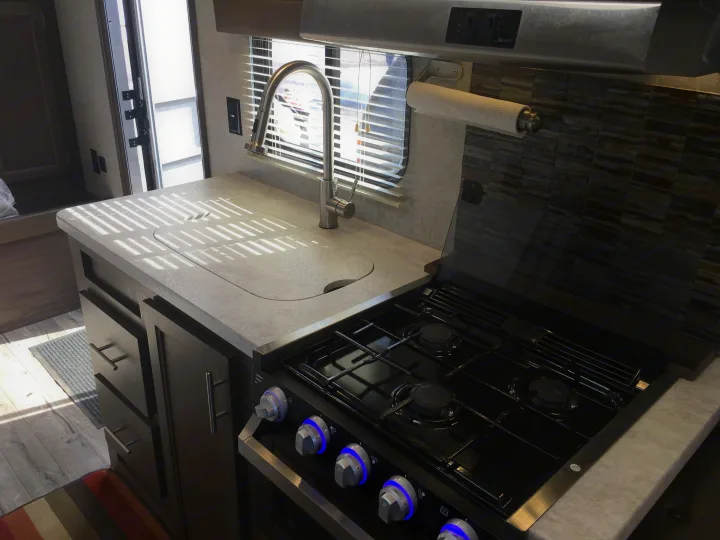 Modern RV rental kitchen featuring a light-colored countertop, stainless steel faucet, four-burner stove, and dark wood cabinets.. Forest River Cherokee Grey Wolf 2018