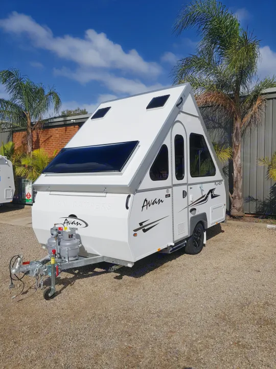 This sleek white A-frame Avan camper, an ideal RV rental option, sits on a gravel lot under a blue sky.. Avan Aliner Camper 2022