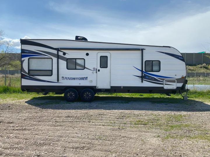 Parked Sandstorm travel trailer RV rental, white with blue graphics, on a gravel and grass lot.. Forest River Sandstorm 2017
