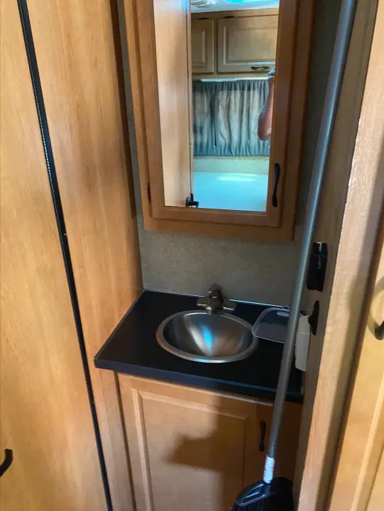 Inside an RV rental, a compact bathroom: stainless steel sink, wood cabinetry, mirror, and a broom leaning nearby.. Thor Motor Coach Four Winds Majestic 2015