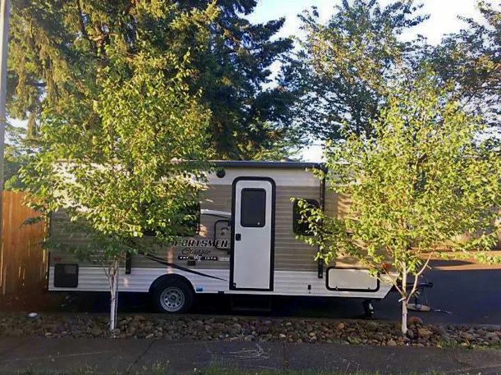 A small Sportsmen Classic RV rental is parked outdoors, partially hidden by two leafy trees, ready for adventure.. KZ SPORTSMEN 2017