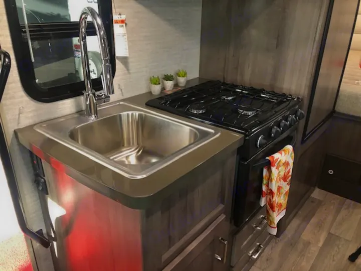 View of a compact RV rental kitchen featuring a stainless steel sink with a modern faucet, a black gas stove/oven, and storage cabinets.. Winnebago Minnie Winnie 2020