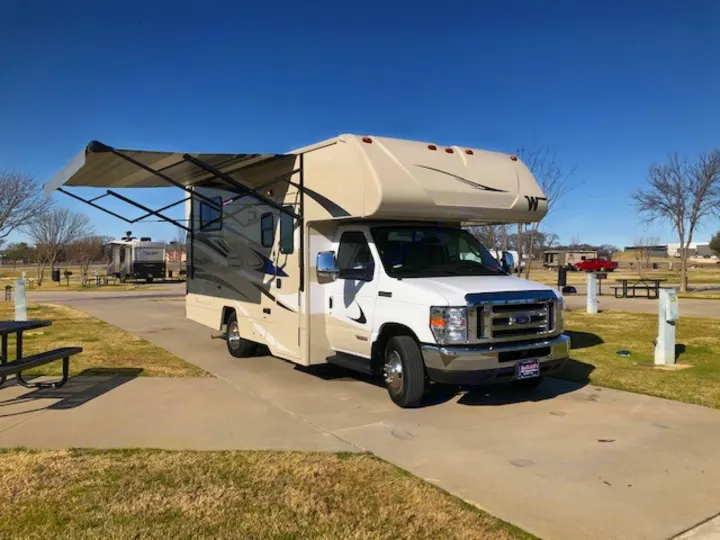 This white and beige RV rental, with its awning extended, is parked on a concrete pad at a sunny campground site.. Winnebago Minnie Winnie 2020