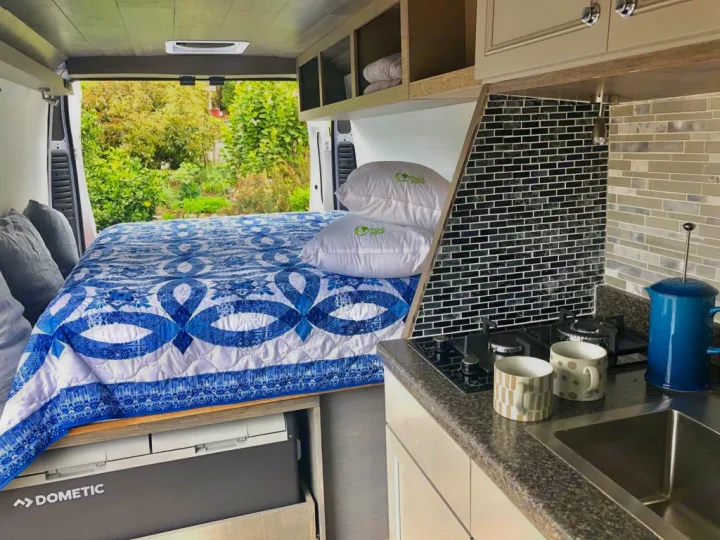 Modern RV rental interior with comfy bed, compact kitchen, and nature views from open rear doors.. Dodge Sprinter 2500 170ext 2015