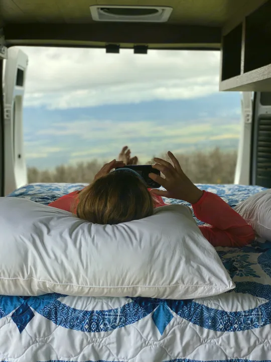 Enjoying comfort in an RV rental, a person relaxes on a bed, phone in hand, admiring the scenic view.. Dodge Sprinter 2500 170ext 2015