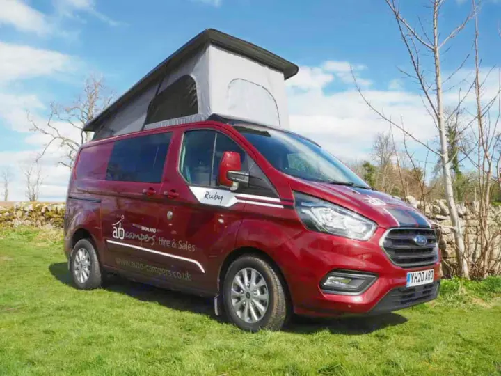 Red pop-top campervan, ideal for an RV rental, parked on green grass under a bright blue sky with trees and a stone wall.. Ford Transit Custom 2+2 berth Pop Top, manual 2020
