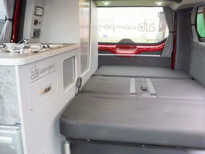 Inside a compact RV rental campervan: a gray fold-out bed fills the rear, with a small kitchen unit visible on the left.. Ford Transit Custom 2+2 berth Pop Top, manual 2020
