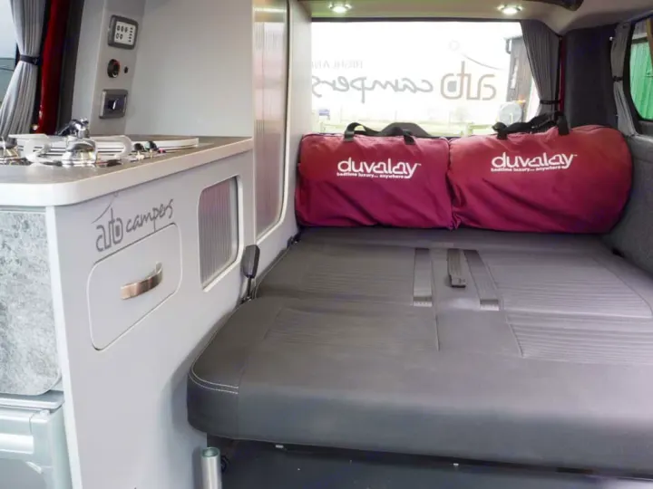 Inside a compact RV rental: kitchenette, pull-out bed with Duvalay bags. 'Auto campers' branding for your travel adventures.. Ford Transit Custom 2+2 berth Pop Top, Manual 2021
