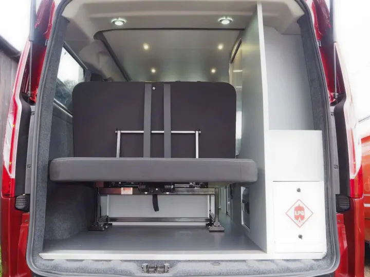 Inside this RV rental, a versatile campervan interior features a dark gray convertible bench/bed, white LPG cabinet, and ceiling lights.. Ford Transit Custom 2+2 berth Pop Top, Manual 2021