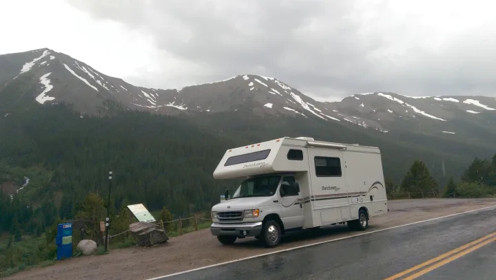 An RV rental parked on a wet mountain road, overlooking snowy peaks & lush forests under a cloudy sky. Perfect scenic road trip.. Dutchmen Other 2001