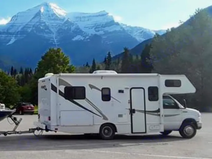 White RV rental with a trailer, parked at a scenic stop overlooking majestic snow-capped mountains and green forests, perfect for nature travel.. Thor Motor Coach Four Winds Majestic 2013