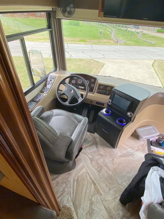 Inside an RV rental: detailed view of the driver's cockpit with dashboard, steering wheel, and front window revealing a paved area.. Coachmen Sportscoach 2018