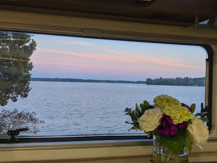 View from an RV rental: a clear window frames a calm lake at sunset. A vase of fresh flowers sits on the counter inside.. Winnebago Navion 24D 2020