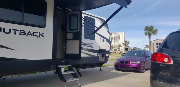 RV rental setup at a sunny campground, with a purple sports car and a dark SUV parked nearby. Perfect for a beach getaway.. Keystone Outback 2020