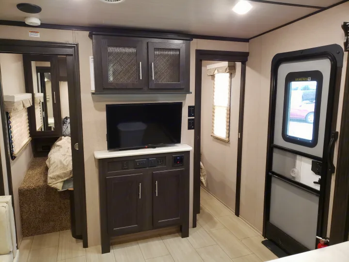 Modern RV rental interior showing living area with TV, dark cabinetry, light flooring. Leads to bedroom & main entrance.. Forest River XLR hyperlite 2019