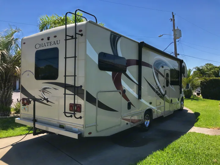 Sunny day view of a light-colored Chateau RV rental with a ladder and bike rack, parked on a driveway with palm trees.. Thor Chateau 2020