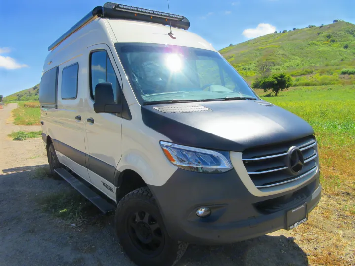 White Winnebago RV rental camper van with off-road tires and a light bar, parked on a sunny dirt trail next to a green hill.. Winnebago Revel 2020