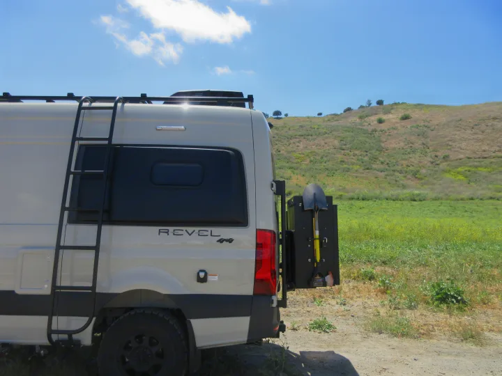 This Winnebago Revel 4x4 camper van, perfect for rv rental adventures, is parked by a grassy hill, ready for off-road exploration with its ladder and shovel.. Winnebago Revel 2020