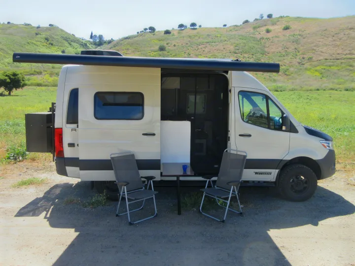 RV rental camper van with side door open, awning extended, and outdoor chairs/table set up on gravel in hilly landscape.. Winnebago Revel 2020