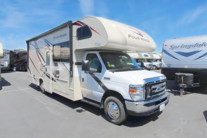 A sleek beige and white Four Winds Class C RV ready for an rv rental journey. Parked on asphalt under a clear blue sky.. Thor Motor Coach Four Winds 2020