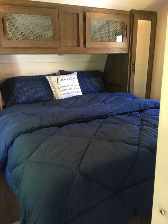 Inside an RV rental bedroom features a bed with blue bedding, a decorative pillow, and light wood cabinets with patterned glass.. Forest River Salem 2021