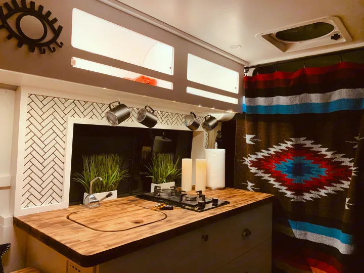 Stylish RV rental kitchen featuring a wood counter, sink, two-burner stove, and a vibrant patterned blanket.. Ram Promaster 2020