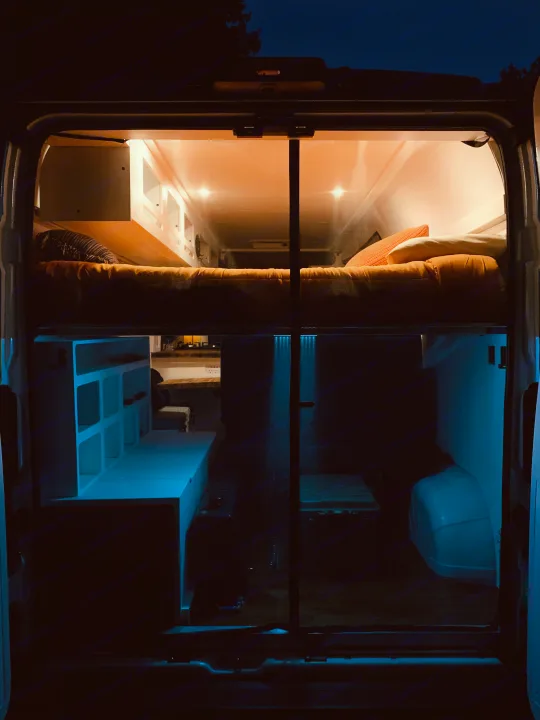 Cozy RV rental interior at night, showing a top bed with warm orange lights and cool blue-lit storage below.. Ram Promaster 2020